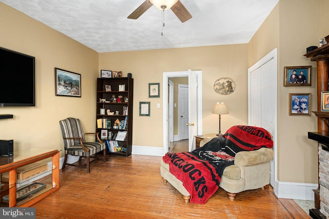 sitting room featuring wood-type flooring, a textured ceiling, a stone fireplace, and ceiling fan