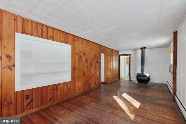 empty room with a wood stove, wooden walls, a baseboard radiator, and dark hardwood / wood-style floors