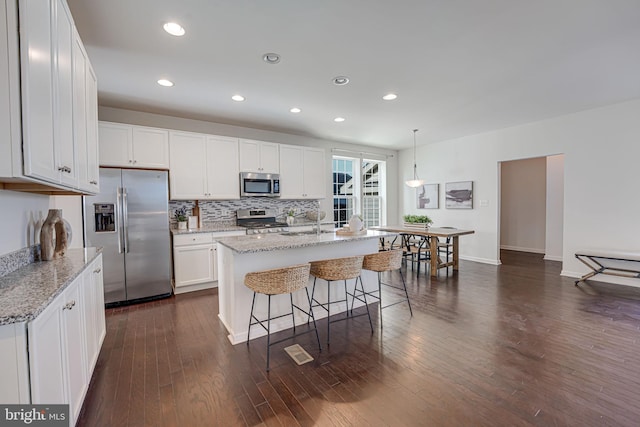 kitchen with pendant lighting, appliances with stainless steel finishes, white cabinetry, a kitchen island with sink, and light stone countertops