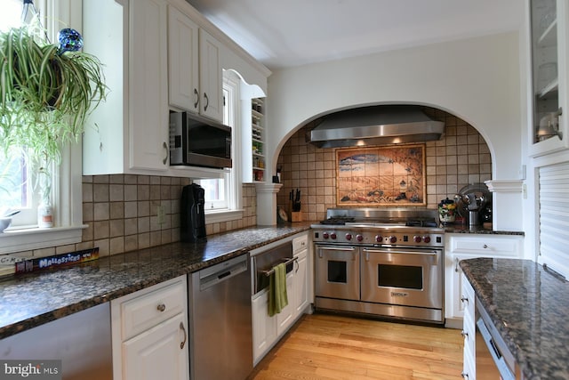 kitchen featuring white cabinetry, wall chimney range hood, dark stone counters, decorative backsplash, and appliances with stainless steel finishes