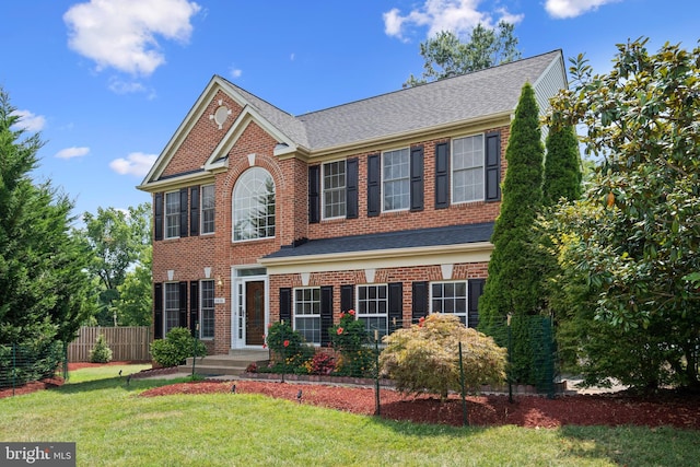 colonial home with brick siding, fence, a front lawn, and roof with shingles