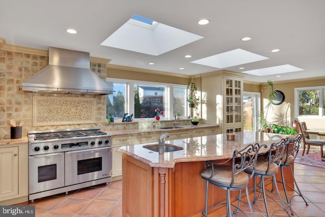 kitchen featuring sink, exhaust hood, light stone countertops, a kitchen island with sink, and range with two ovens