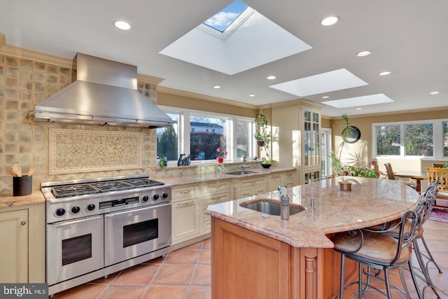 kitchen with a kitchen island with sink, double oven range, light stone countertops, range hood, and a breakfast bar area