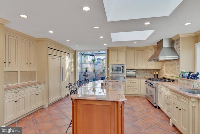 kitchen featuring cream cabinets, a kitchen island with sink, built in appliances, light stone countertops, and wall chimney range hood