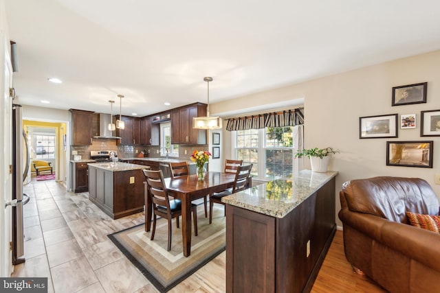 kitchen featuring decorative light fixtures, tasteful backsplash, light stone countertops, dark brown cabinets, and wall chimney range hood