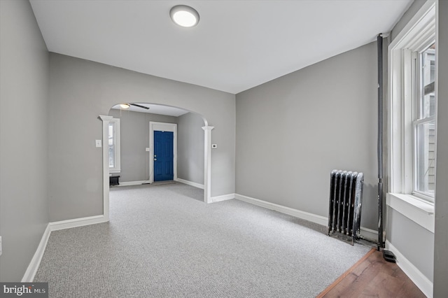 carpeted empty room with arched walkways, baseboards, ceiling fan, and radiator heating unit