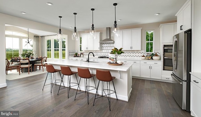 kitchen with white cabinetry, wall chimney exhaust hood, an island with sink, and appliances with stainless steel finishes