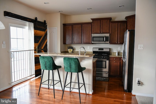 kitchen with light stone countertops, appliances with stainless steel finishes, a kitchen breakfast bar, dark wood-type flooring, and an island with sink