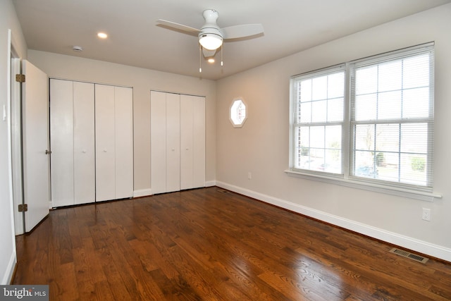 unfurnished bedroom with multiple windows, ceiling fan, dark wood-type flooring, and multiple closets