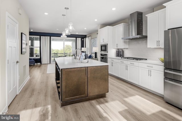 kitchen with appliances with stainless steel finishes, wall chimney exhaust hood, decorative light fixtures, white cabinets, and an island with sink