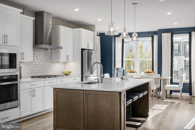 kitchen with white cabinetry, wall chimney exhaust hood, an island with sink, and appliances with stainless steel finishes