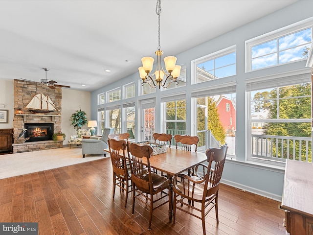 dining room with ceiling fan with notable chandelier, a stone fireplace, and dark wood-type flooring