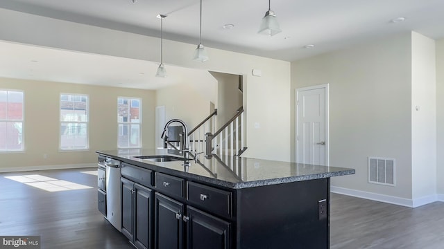 kitchen with sink, dark hardwood / wood-style floors, dark stone counters, hanging light fixtures, and a kitchen island with sink