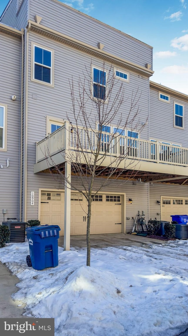 snow covered house featuring cooling unit, a balcony, and a garage