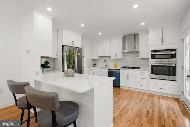 kitchen featuring wall chimney exhaust hood, a kitchen breakfast bar, kitchen peninsula, white cabinets, and appliances with stainless steel finishes