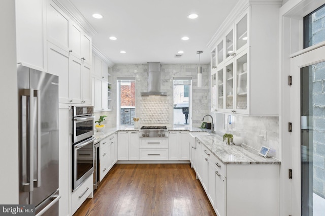 kitchen featuring appliances with stainless steel finishes, light stone countertops, white cabinets, wall chimney range hood, and decorative light fixtures