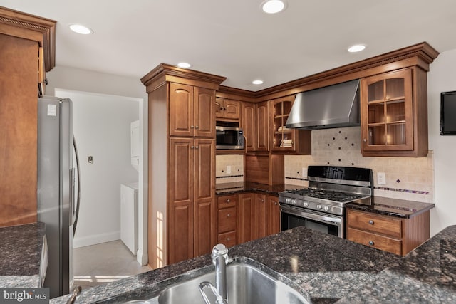 kitchen with stacked washer and clothes dryer, brown cabinets, stainless steel appliances, glass insert cabinets, and wall chimney range hood