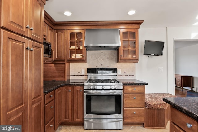 kitchen featuring appliances with stainless steel finishes, wall chimney exhaust hood, tasteful backsplash, dark stone countertops, and glass insert cabinets