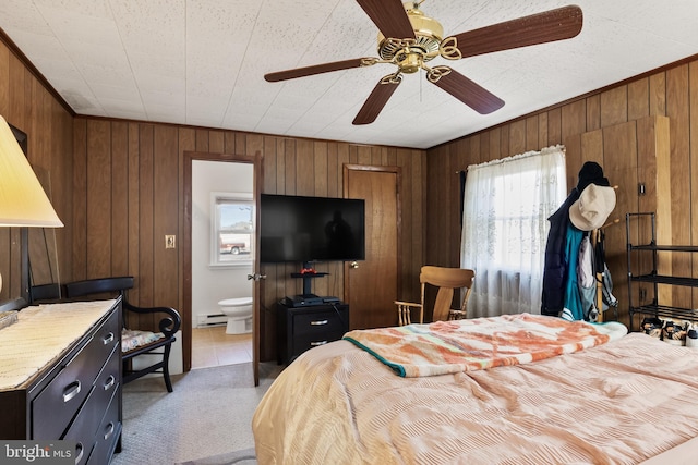 carpeted bedroom featuring baseboard heating, ensuite bathroom, ornamental molding, ceiling fan, and wooden walls