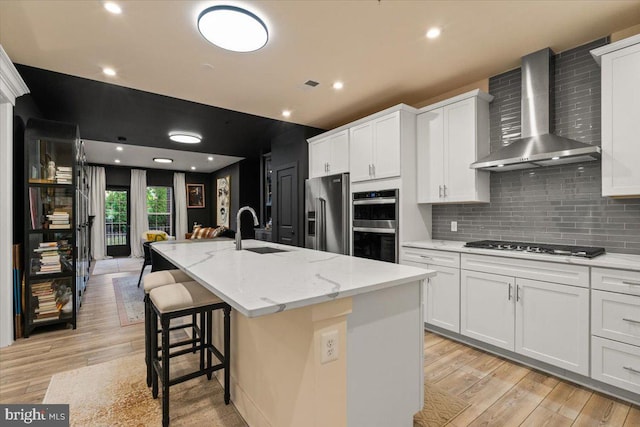 kitchen with white cabinets, stainless steel appliances, wall chimney exhaust hood, and an island with sink