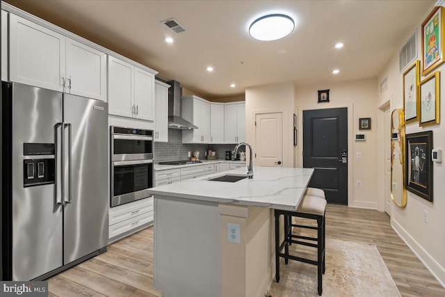 kitchen featuring light stone countertops, sink, wall chimney range hood, an island with sink, and appliances with stainless steel finishes