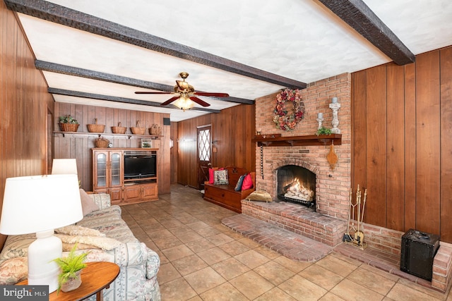 living room featuring ceiling fan, beamed ceiling, wooden walls, a fireplace, and light tile patterned floors