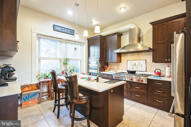 kitchen featuring a breakfast bar, a center island with sink, wall chimney exhaust hood, decorative light fixtures, and stainless steel appliances