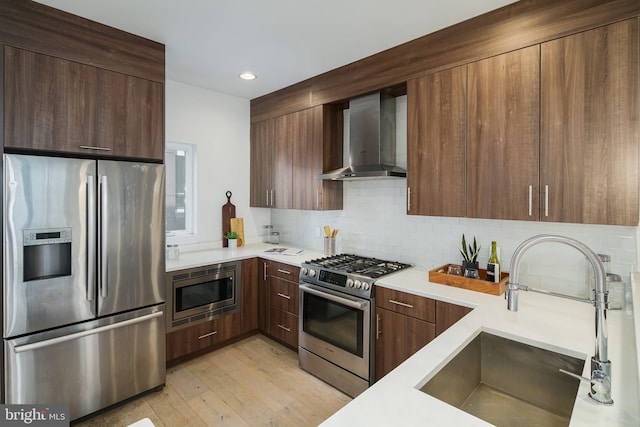 kitchen featuring backsplash, wall chimney range hood, sink, light hardwood / wood-style flooring, and appliances with stainless steel finishes