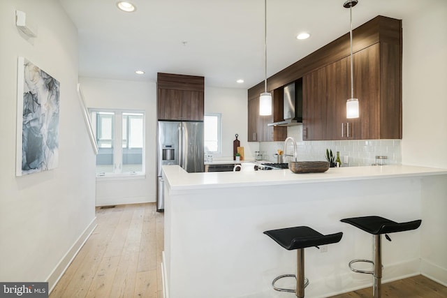 kitchen with kitchen peninsula, a kitchen bar, wall chimney range hood, decorative light fixtures, and stainless steel fridge with ice dispenser