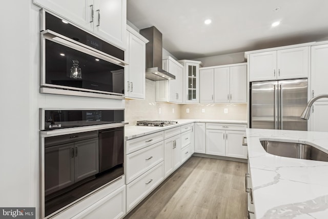 kitchen with light stone counters, stainless steel appliances, sink, wall chimney range hood, and white cabinetry