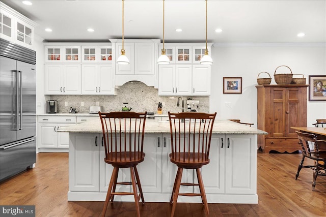 kitchen featuring decorative light fixtures, white cabinetry, an island with sink, stainless steel built in fridge, and light stone counters