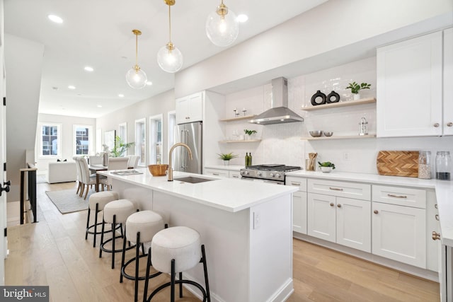 kitchen featuring wall chimney exhaust hood, stainless steel appliances, a kitchen island with sink, pendant lighting, and white cabinetry