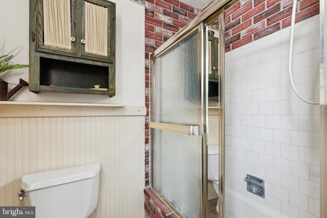 bathroom with combined bath / shower with glass door, toilet, and brick wall