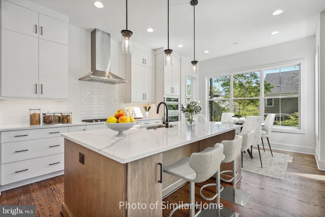 kitchen with wall chimney exhaust hood, white cabinetry, an island with sink, and pendant lighting