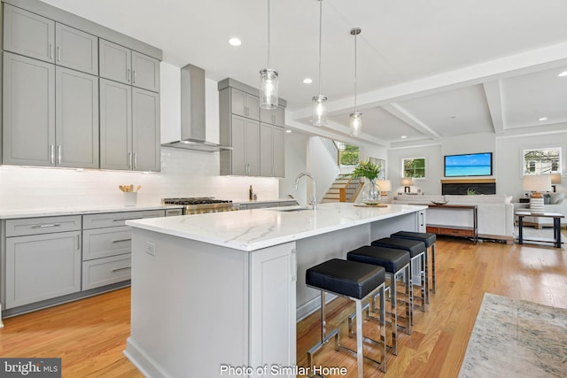 kitchen with gray cabinetry, wall chimney exhaust hood, decorative backsplash, an island with sink, and decorative light fixtures