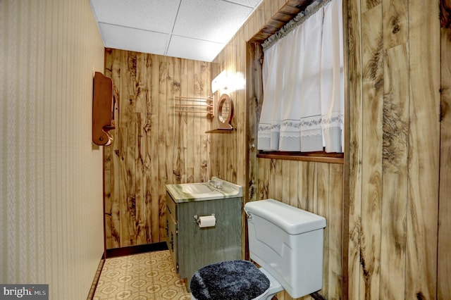 bathroom with wooden walls, vanity, toilet, and a drop ceiling