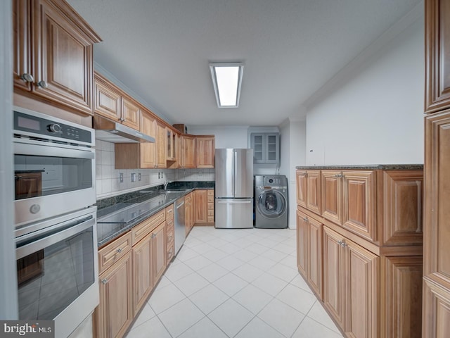 kitchen featuring under cabinet range hood, stainless steel appliances, brown cabinets, dark stone counters, and washer / clothes dryer