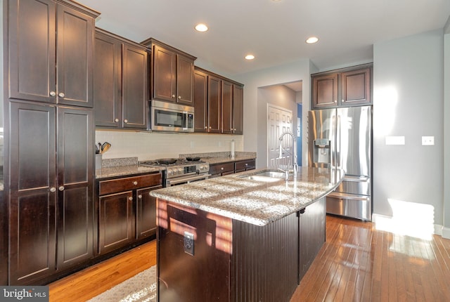 kitchen with sink, light stone countertops, an island with sink, dark brown cabinets, and stainless steel appliances