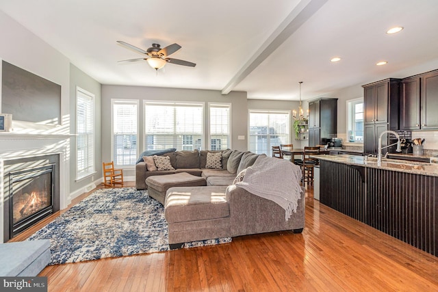 living room featuring ceiling fan with notable chandelier, sink, a fireplace, beam ceiling, and light hardwood / wood-style floors