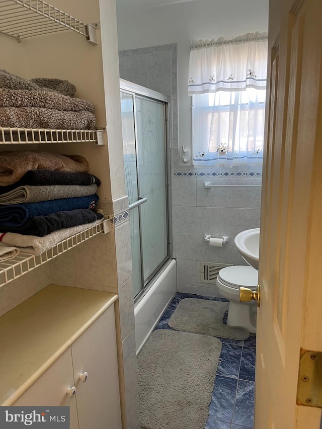 bathroom featuring tile patterned flooring, toilet, bath / shower combo with glass door, and tile walls