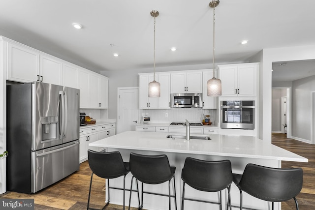 kitchen with stainless steel appliances, a center island with sink, hanging light fixtures, and dark wood-type flooring