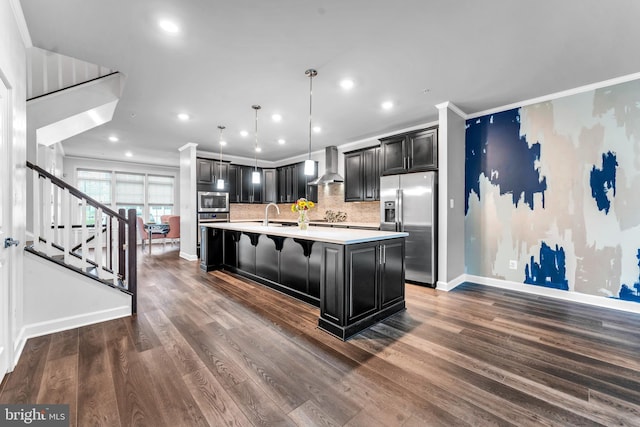 kitchen with appliances with stainless steel finishes, ornamental molding, wall chimney range hood, dark hardwood / wood-style floors, and an island with sink