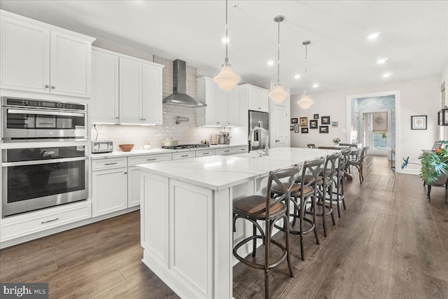 kitchen with pendant lighting, a breakfast bar, a center island with sink, white cabinets, and wall chimney exhaust hood