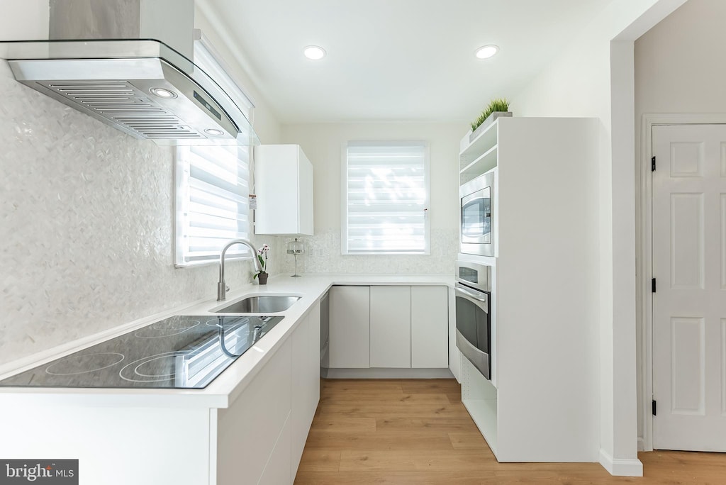 kitchen featuring white cabinets, wall chimney range hood, sink, light wood-type flooring, and appliances with stainless steel finishes