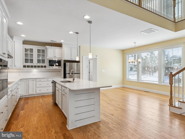 kitchen with appliances with stainless steel finishes, light wood-type flooring, pendant lighting, white cabinets, and an island with sink