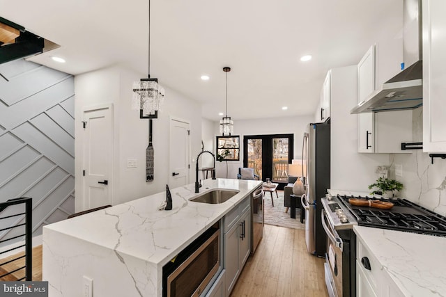 kitchen featuring white cabinetry, sink, stainless steel appliances, wall chimney range hood, and an island with sink