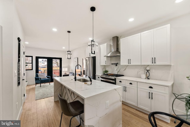 kitchen featuring white cabinetry, light stone countertops, wall chimney range hood, a center island with sink, and appliances with stainless steel finishes