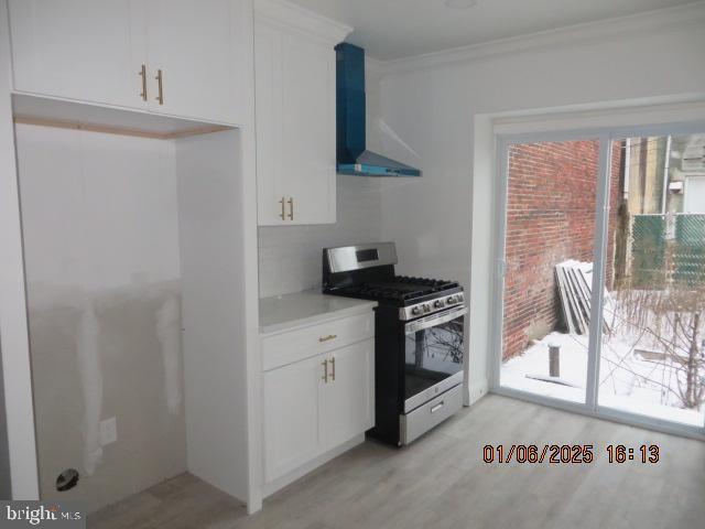 kitchen with wall chimney exhaust hood, ornamental molding, light hardwood / wood-style floors, white cabinetry, and stainless steel range with gas stovetop