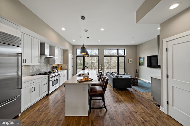 kitchen featuring a kitchen island with sink, wall chimney range hood, premium appliances, white cabinets, and light countertops