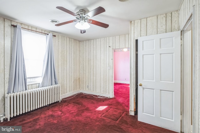 carpeted spare room with wooden walls, radiator heating unit, ceiling fan, and plenty of natural light